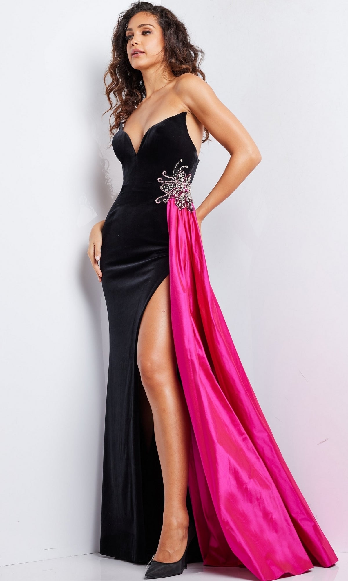 A woman models the Jovani Side-Sash Strapless Long Prom Dress 26117, featuring velvet fabric, a thigh-high slit, an embellished brooch, and a dramatic pink sash, standing against a white background.